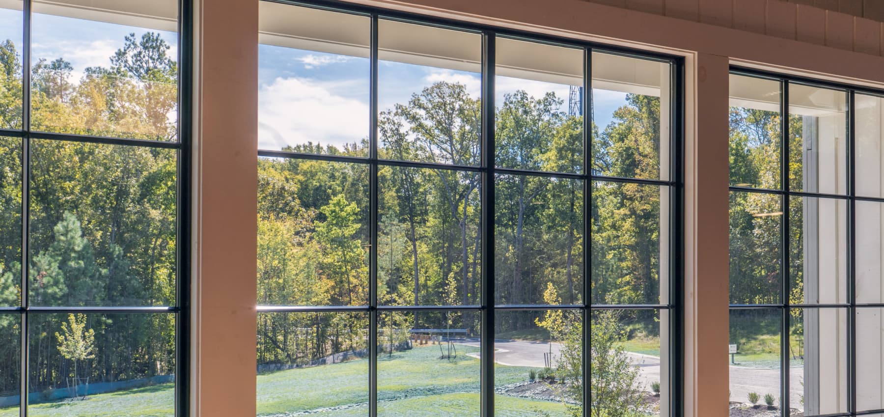 interior-view-of-floor-to-ceiling-black-framed-curtain-wall-windows-overlooking-a-landscaped-lawn-and-tree-line